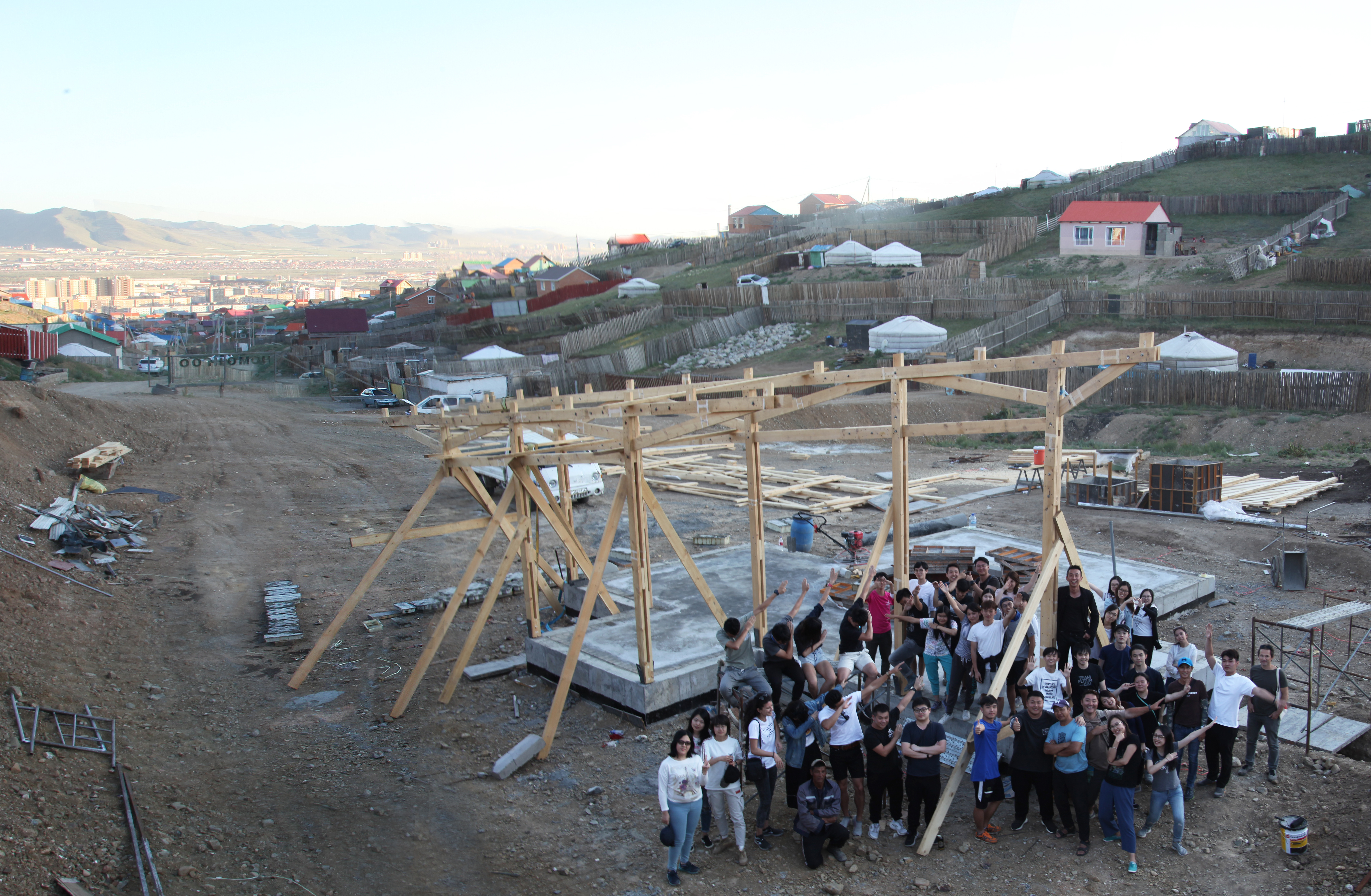  Students amassed in a construction site next to a partially constructed wooden framework. Individual ger plots can be seen on the hills in the distance. 